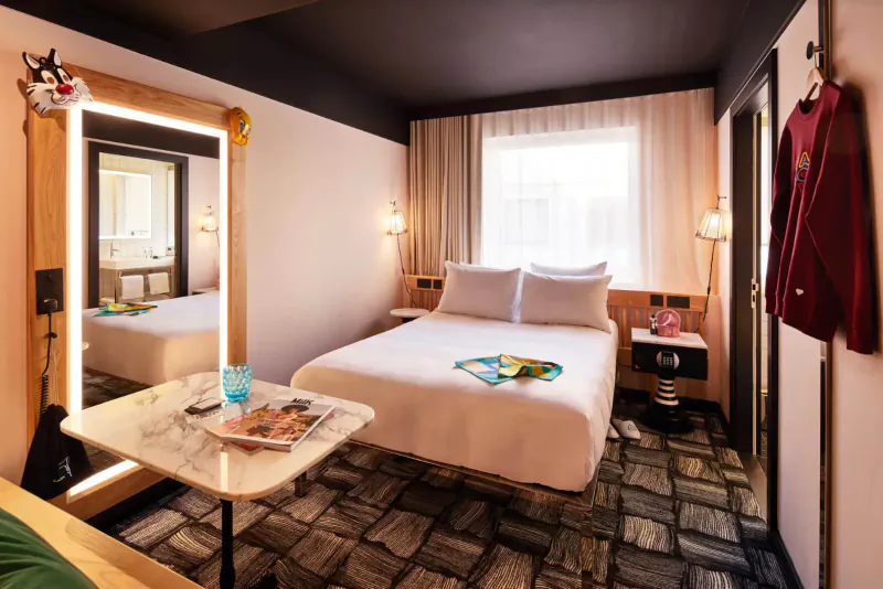 Cozy Mama Shelter hotel room with white bed, magazines, toys on table, red jacket hung, large mirror, and patterned floor.