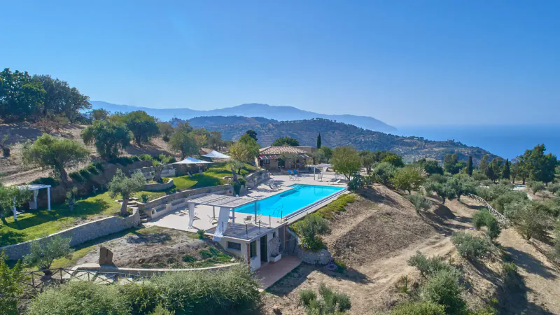 Aerial view of luxury Wish Sicily villa with infinity pool, gardens, and Mediterranean mountains and sea beyond.
