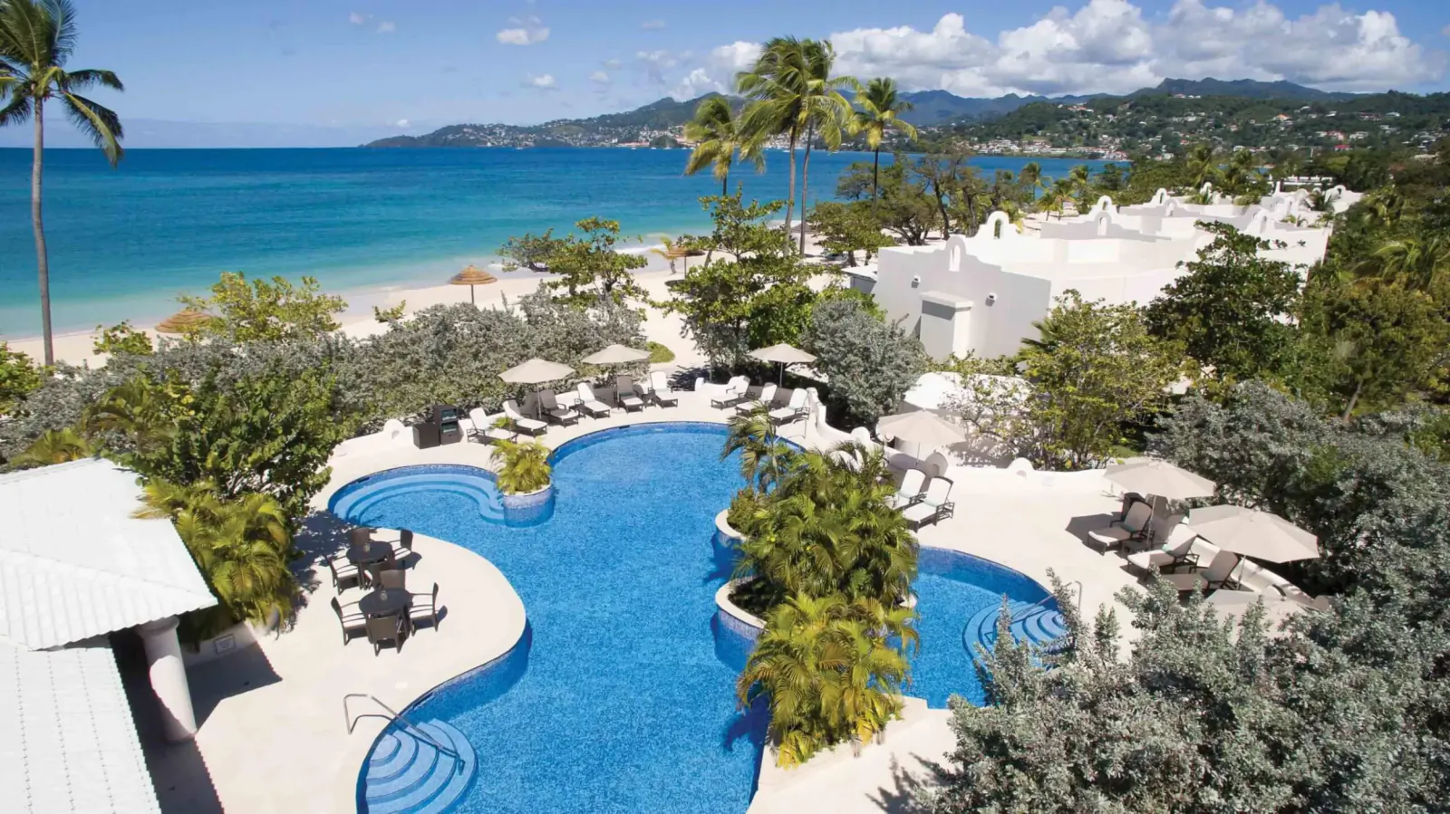 Aerial view of Spice Island Beach Resort's curved turquoise pool, lounge chairs, palms, white buildings, beach, and ocean with mountains in Grenada.
