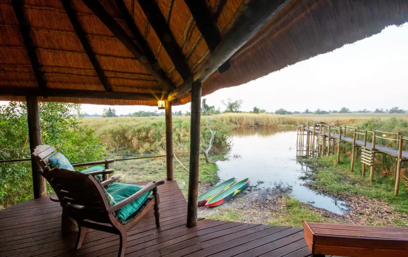 Open-sided thatched lodge deck with green chairs overlooking Okavango Delta marsh, wooden walkway, kayaks, and river at dusk.