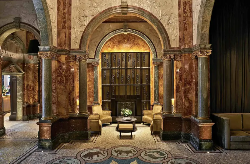 Grand arched lobby of Kimpton Fitzroy London with ornate columns, bear mosaic floor, beige armchairs, and lit fireplace.