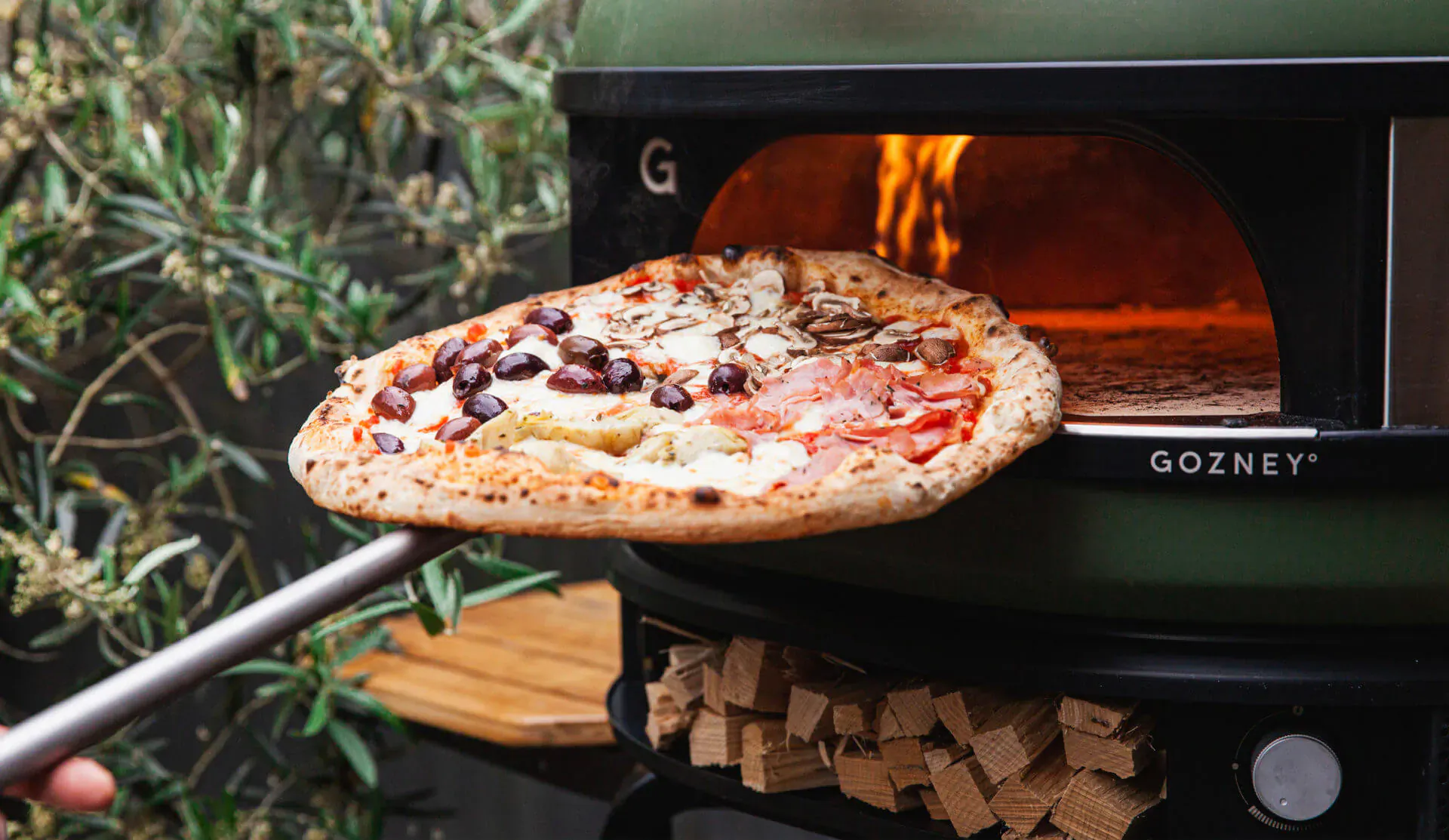 GoZney green wood-fired pizza oven with flames, peel holding topped pizza with olives emerging from open door, garden setting.