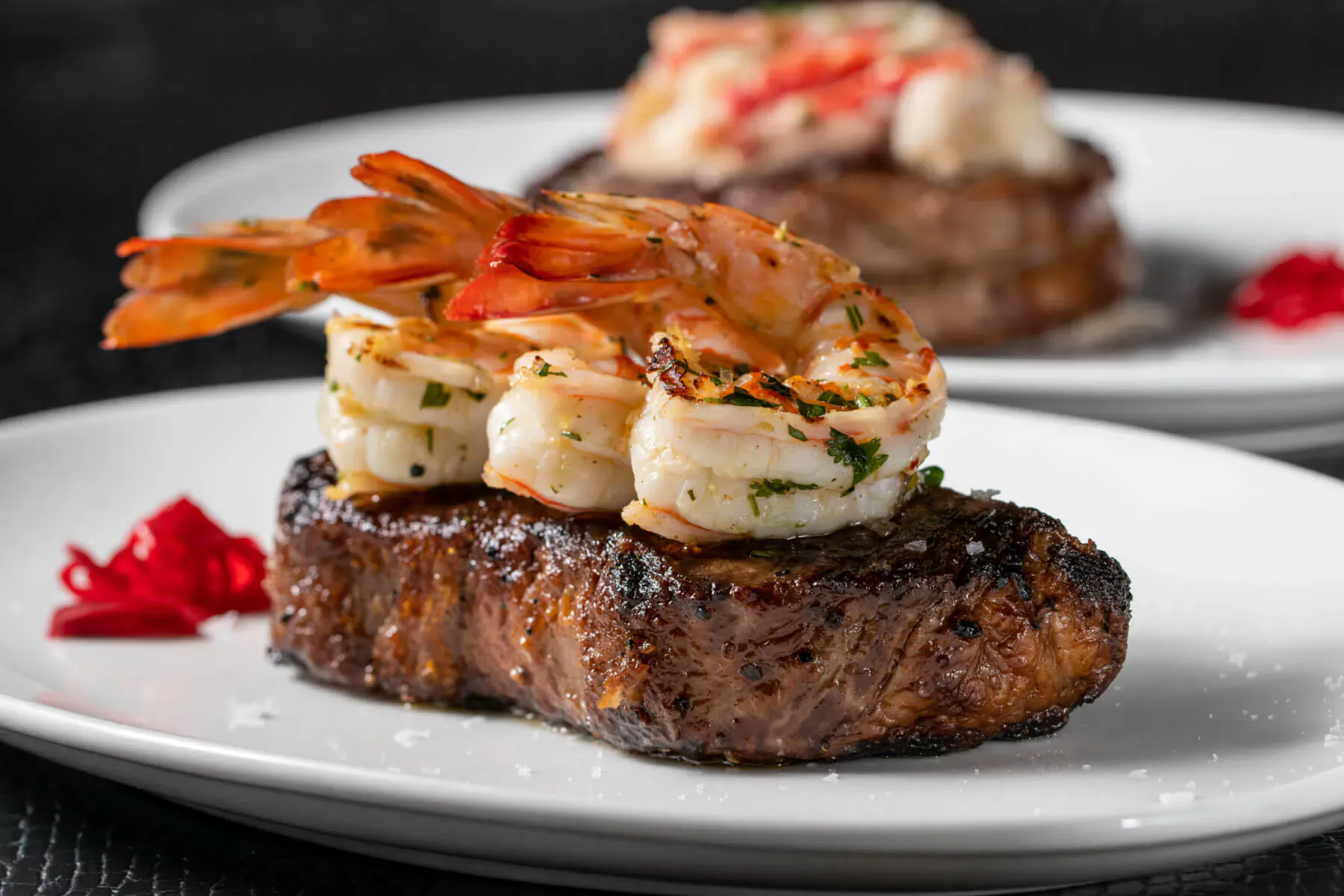 Grilled steak topped with large shrimp, parsley, and red garnishes on white plates at STK.