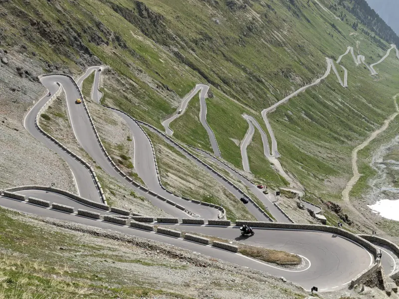 Aerial view of winding mountain pass road with supercars driving on steep green Alpine slopes.