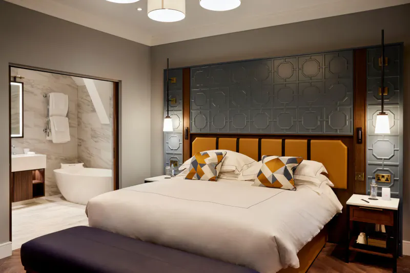 Luxurious hotel bedroom with king bed, geometric headboard, en-suite marble bathroom with freestanding tub.