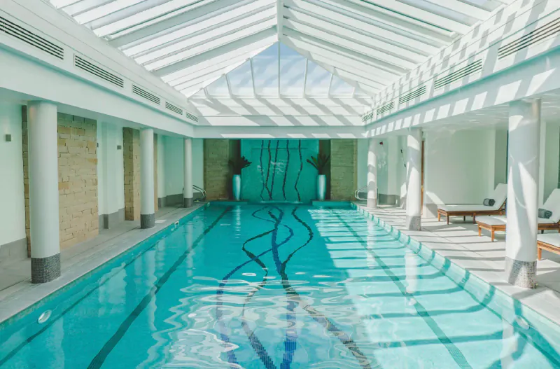 Elegant indoor swimming pool with swirling turquoise water feature, glass atrium ceiling, columns, and lounge chairs in luxury hotel spa.