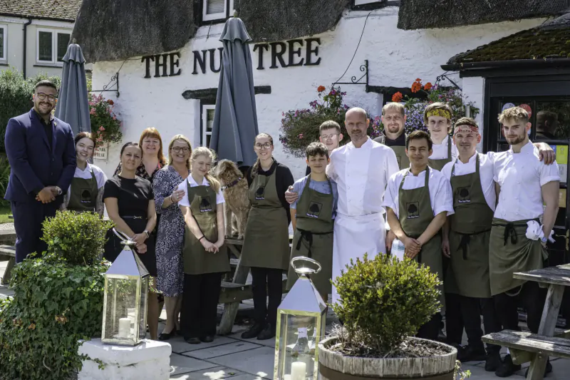 Group of chefs and staff in aprons posing outside The Nut Tree Inn pub, with lanterns and plants.