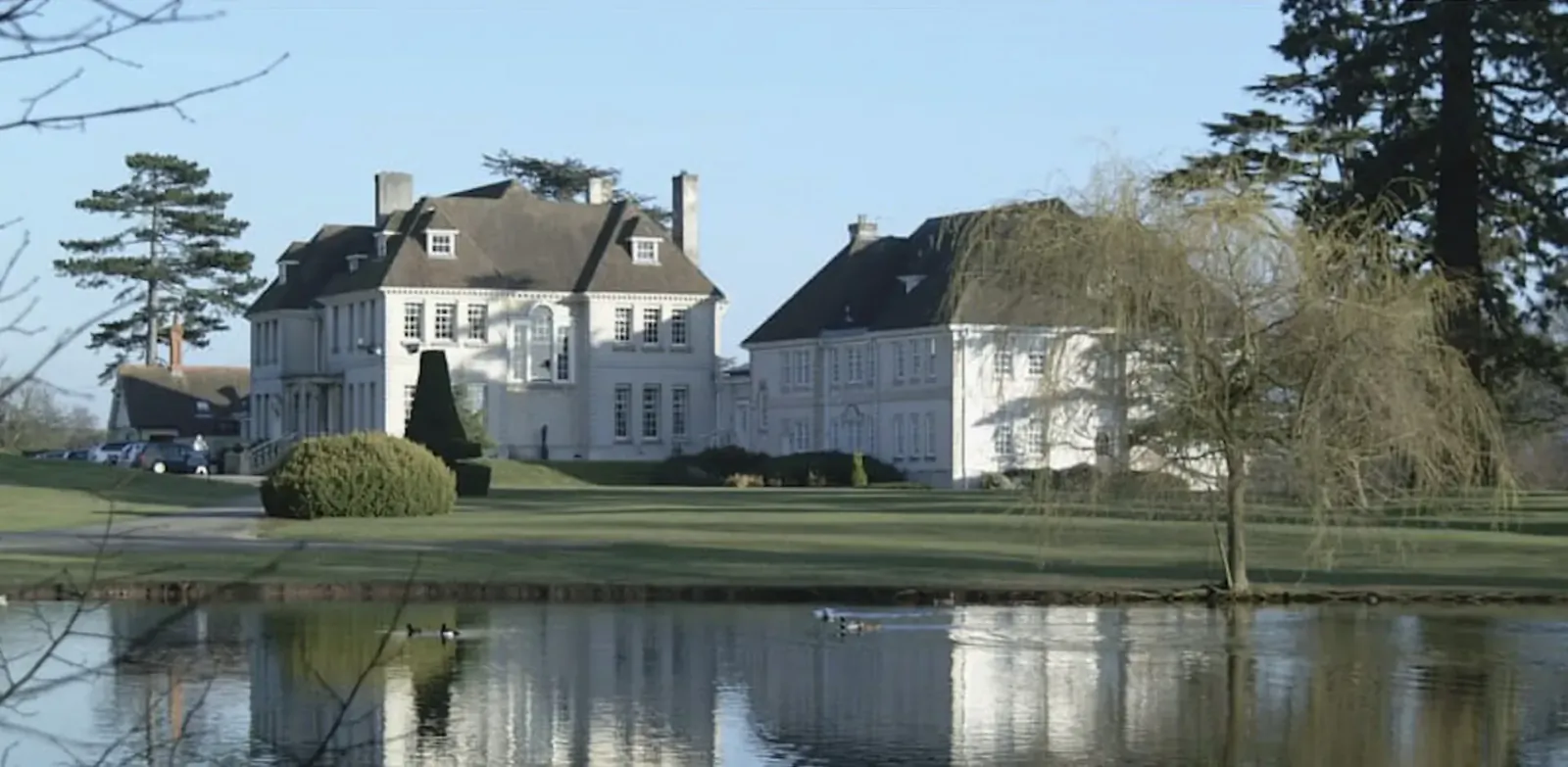 Brockencote Hall luxury mansion with white facade, manicured lawns, tall pines, and pond reflection.