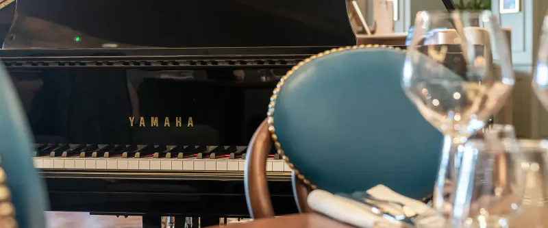 Close-up of black Yamaha grand piano beside turquoise leather chair, wine glasses, and table setting at Browns Brasserie.
