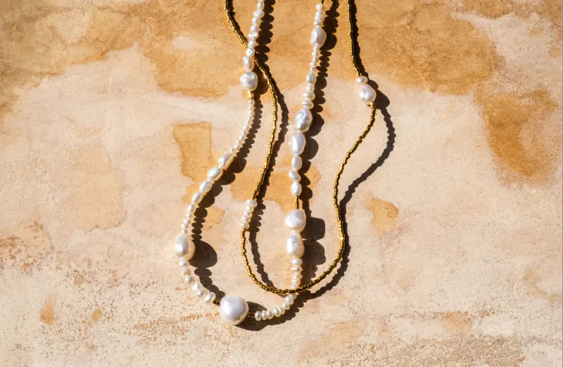 Three pearl necklaces with looped ropes draped on textured beige surface, Pebble & Loop style.