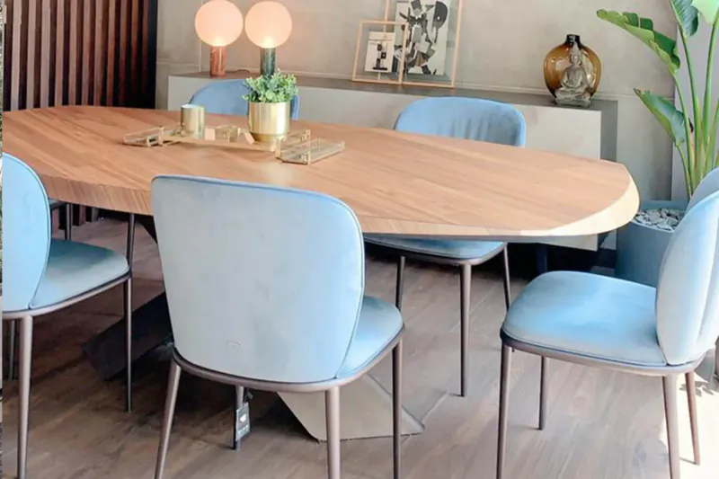 Modern oval wooden dining table with blue velvet chairs, plants, lamps, and framed photos in elegant room
