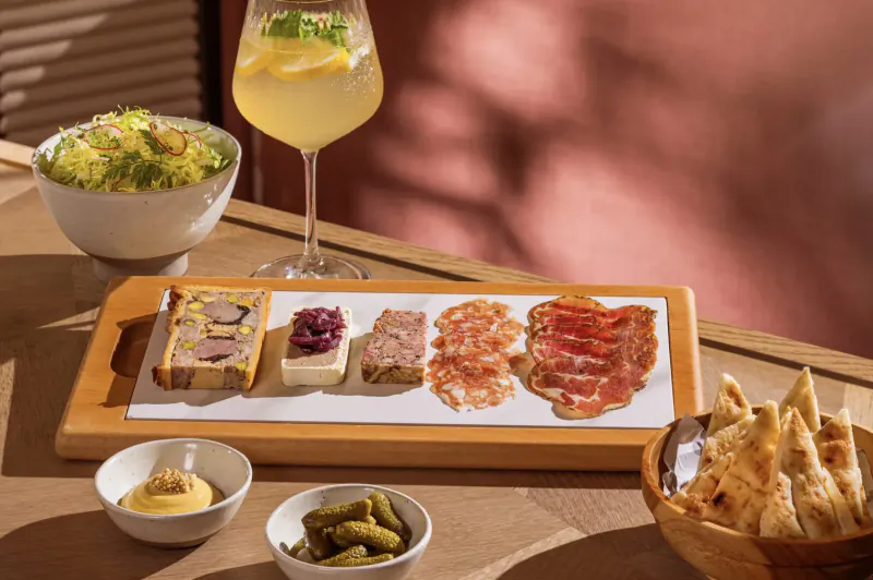 Wooden board with pâté, cheese, cured meats, mustard, pickles; side greens, flatbread, lemon gimlet glass on table.