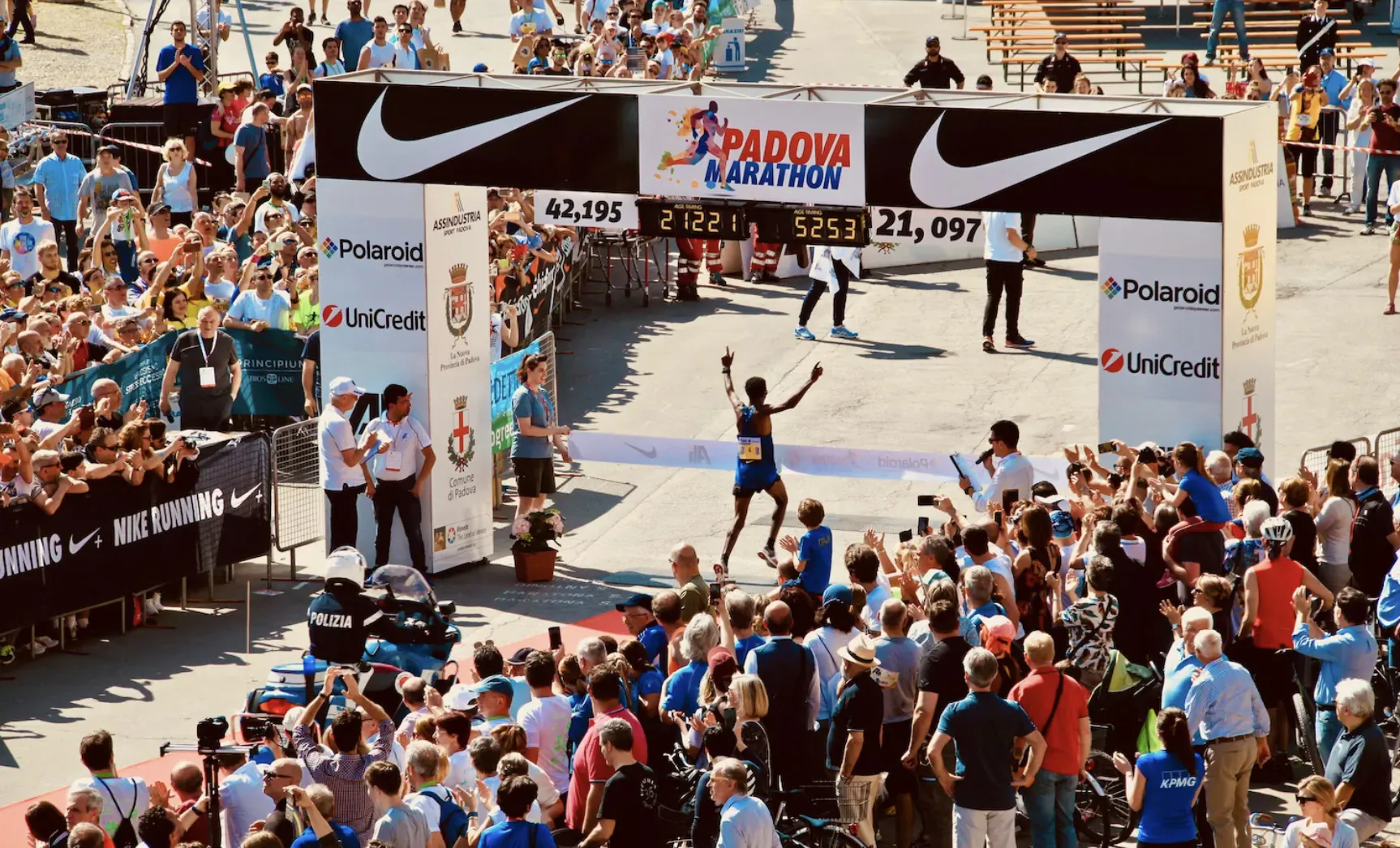 Runner in blue breaks through finish line at 2021 Padova Marathon, Nike arch, cheering crowds.