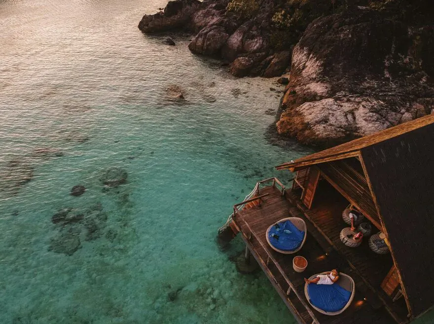 Aerial view of luxurious overwater bungalow with blue loungers on deck, turquoise lagoon, rocks, and sunset sea.
