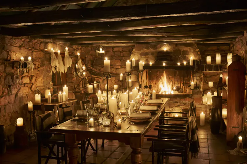 Cozy candlelit stone cellar with wooden dining table set for a meal by a crackling fireplace at Bushmans Kloof retreat.