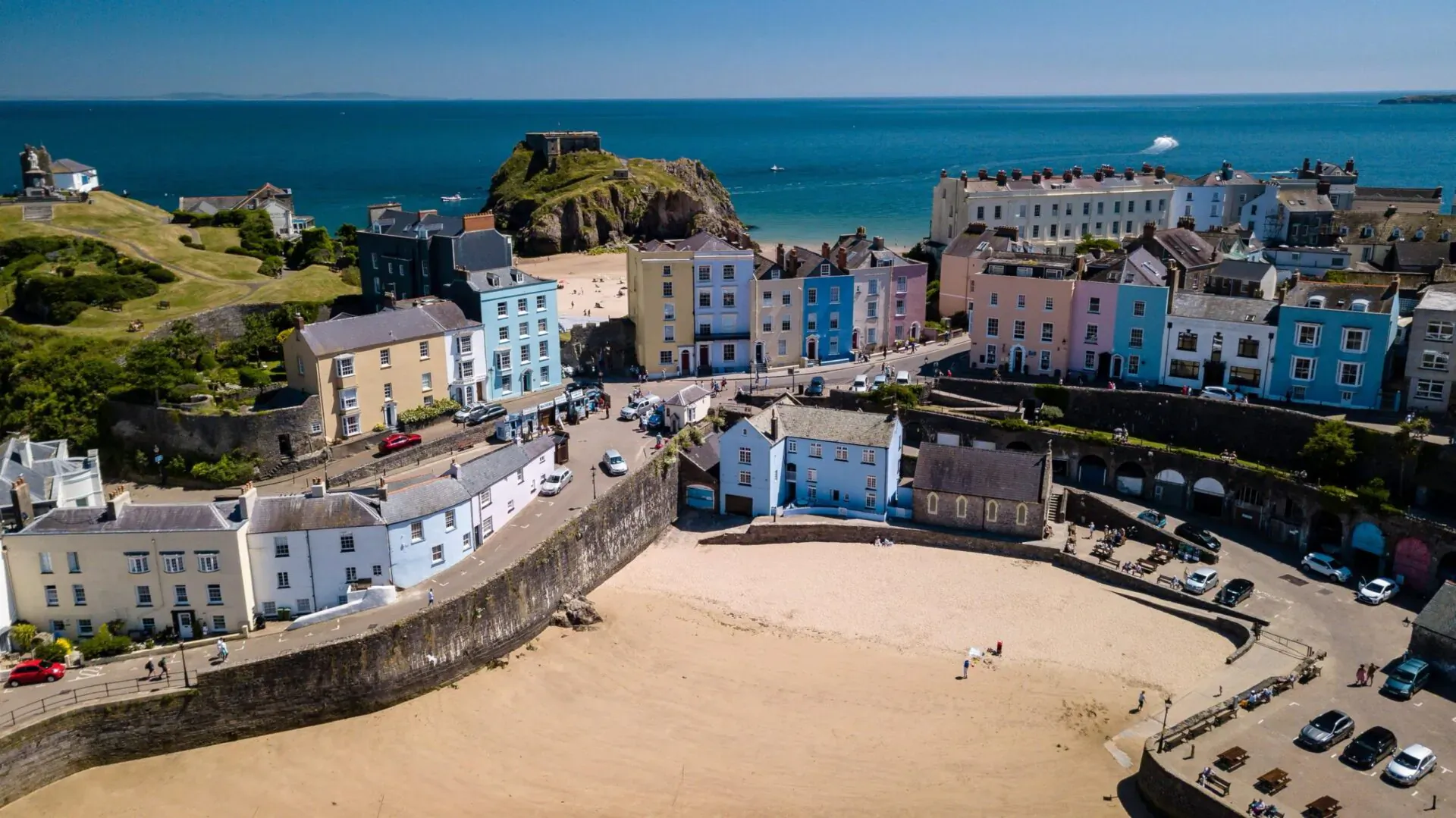 Aerial view of colorful terraced houses surrounding a sandy bay in Tenby, with sea and cliffs beyond.