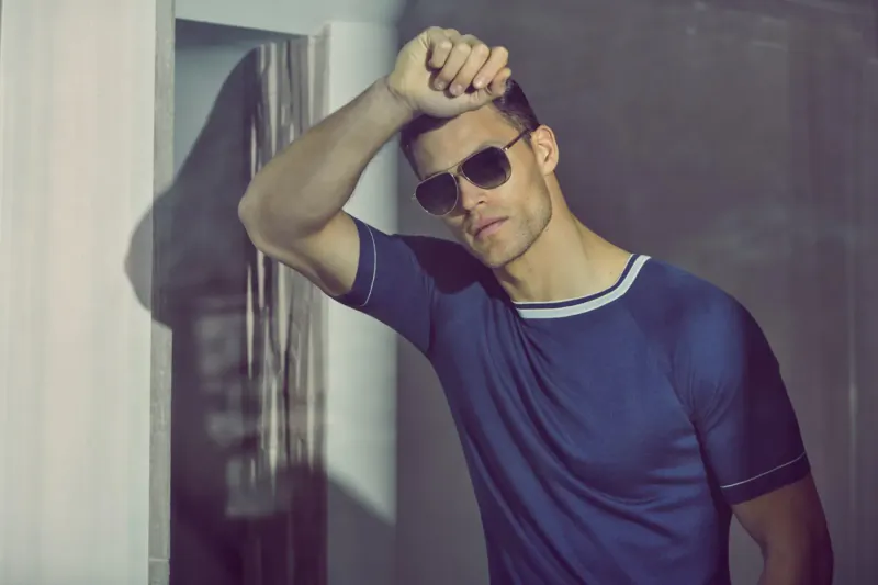 Man in sunglasses and navy short-sleeve shirt poses with hand on head against doorframe, shadowy interior.