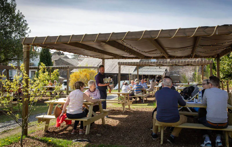 Group of people seated at picnic tables under shaded pergola in garden at The Black Bull, one woman holds flowers
