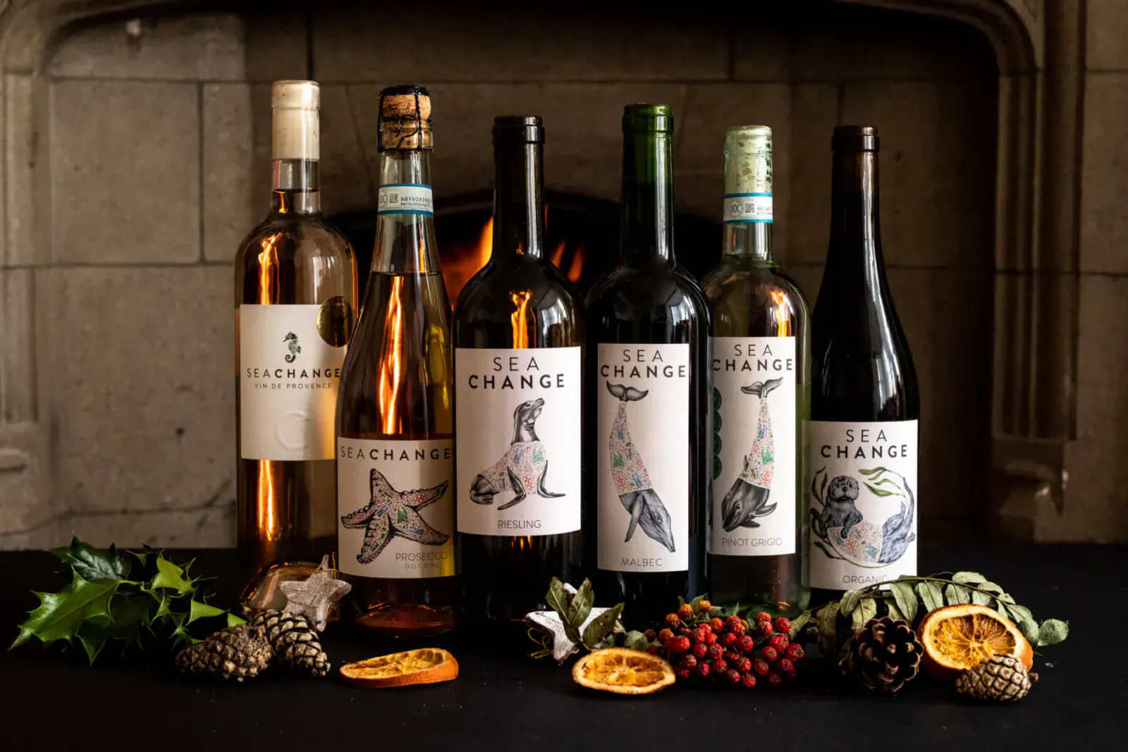 Sea Change eco-wines: rosé, white, red bottles with sea creature labels on table with holly, pinecones, oranges by stone fireplace.