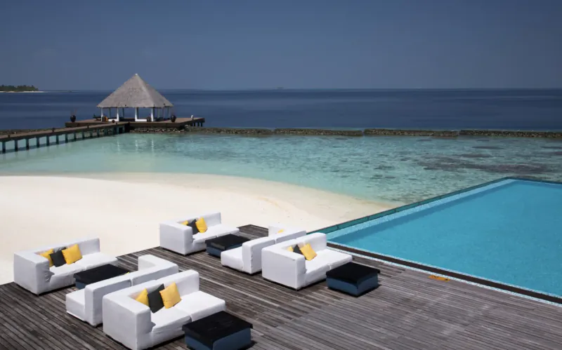 Luxury white outdoor sofas with yellow pillows on wooden deck beside infinity pool, white sand beach, turquoise sea, and thatched pavilion at Maldivian resort