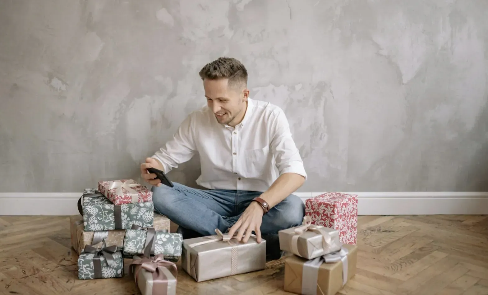 Smiling man in white shirt and jeans sitting on floor, selecting from stack of wrapped gifts he'll love.