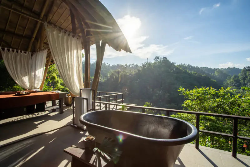 Luxury open-air spa balcony at Buahan Bali with copper bathtub, massage table, and lush jungle valley view under sunny sky.