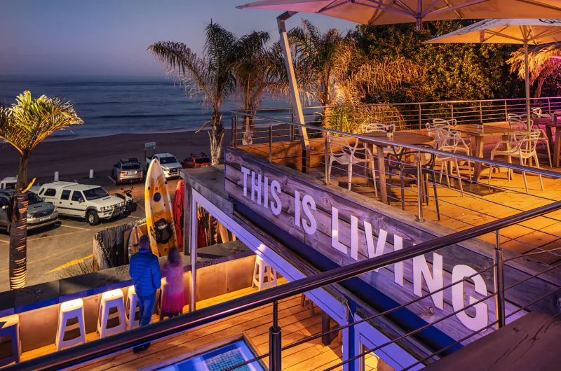Twilight view of The Bungalow seaside deck in Plettenberg Bay with 'THIS IS LIVING' sign, palm trees, ocean, cars, and surfers.