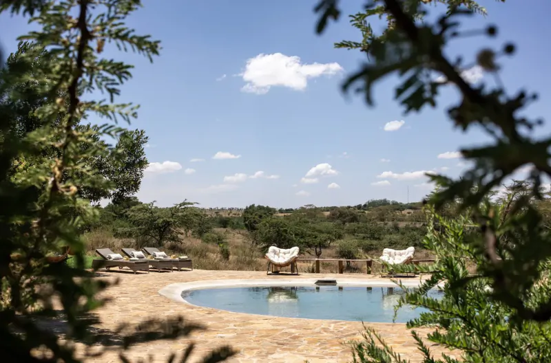 Framed by acacia branches, oval turquoise pool with white hanging chairs at El Karama Lodge in savanna landscape under blue sky.
