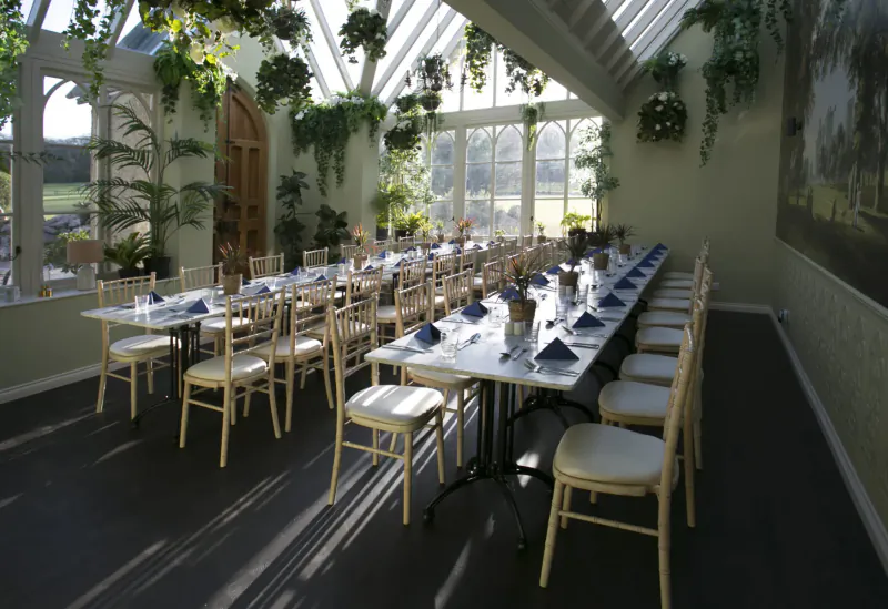 Elegant conservatory dining room at Parc le Breos with long white table set for banquet, blue napkins, wooden chairs, plants, and skylights.