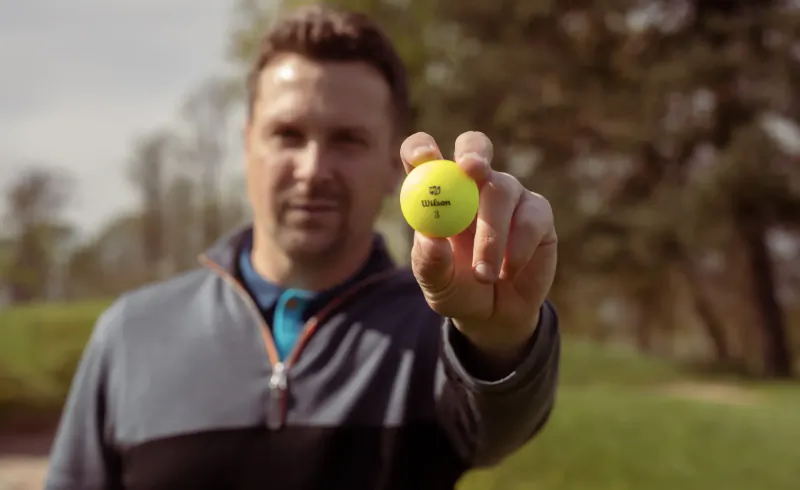 Man in golf attire holding up yellow Wilson golf ball on course, Valentine's gift idea