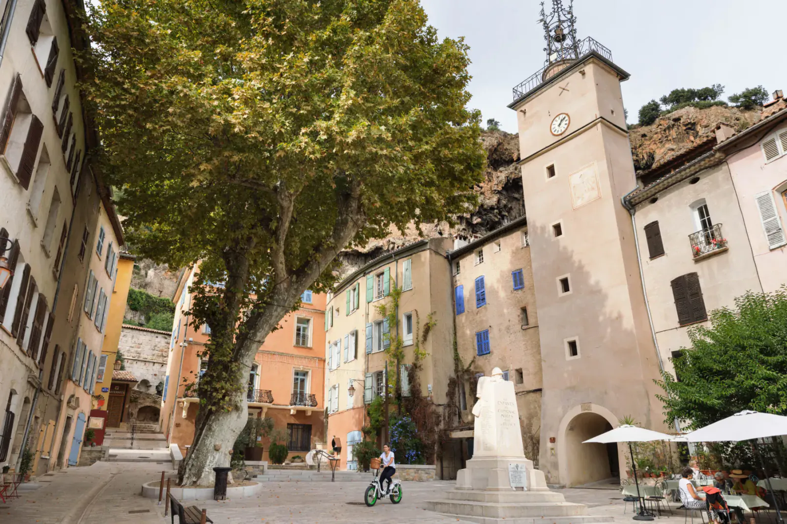 Sunny Provençal square with colorful shuttered buildings, tall clock tower, large tree, statue, and people biking near café umbrellas