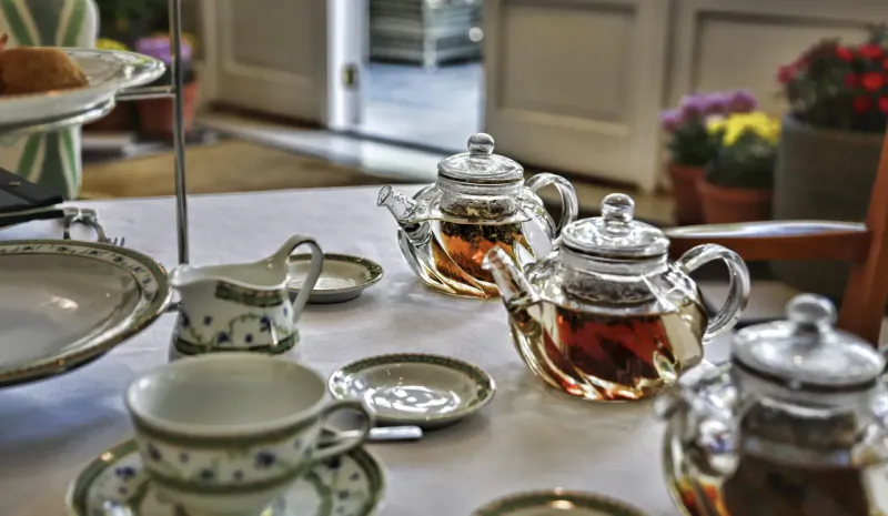 Elegant afternoon tea setup with glass teapots of amber tea, floral cups, scones on tiered stand amid potted roses at Mount Nelson.