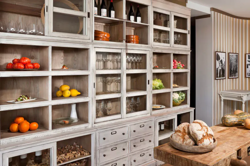 White wooden kitchen cabinetry stocked with glassware, wine bottles, oranges, tomatoes, lemons, bread, and vegetables on a wooden counter.