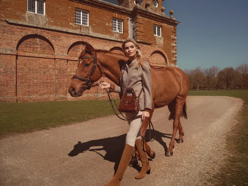 Blonde woman in gray suit and boots leads brown horse on path by brick manor house