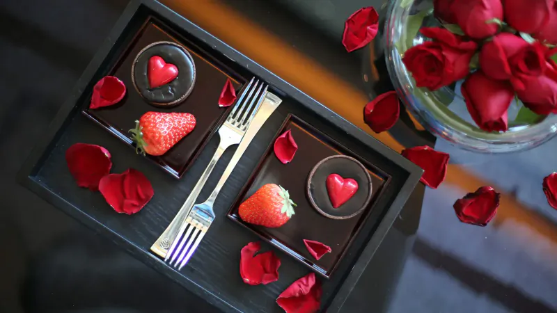 Romantic tray with heart-shaped chocolates, strawberries, forks, and scattered red rose petals beside a vase of roses.