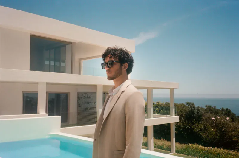 Man in beige suit and sunglasses stands by infinity pool at modern white seaside house under blue sky