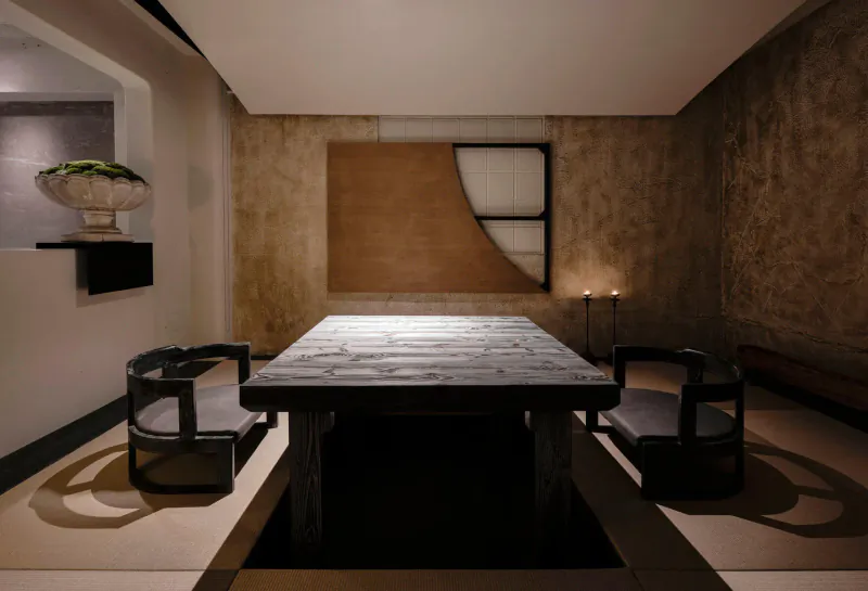 Stylish contemporary restaurant interior with rustic wood dining table, black chairs, beige textured walls, abstract art, plant, and candles.