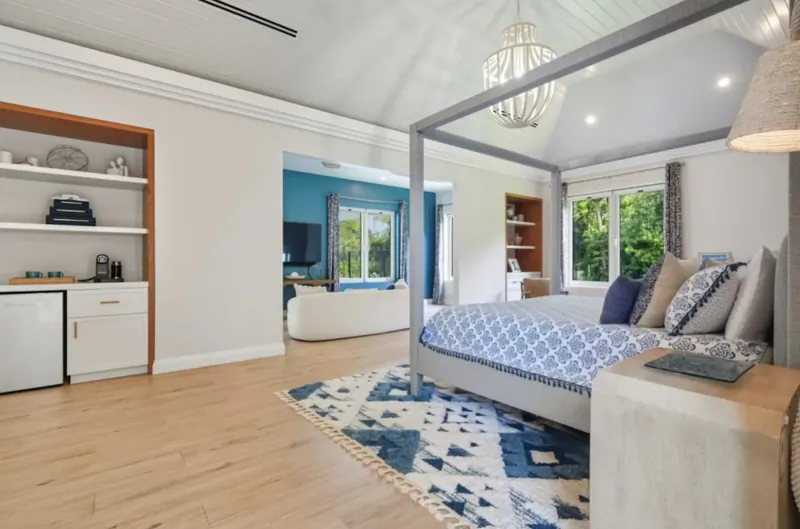Spacious Lyford Cay bedroom with gray four-poster bed, blue duvet, ensuite bath, hardwood floors, and rattan chandelier.