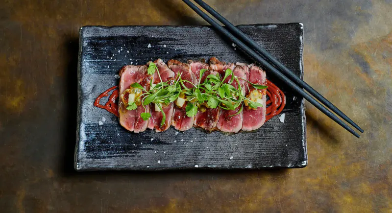 Sliced tuna sashimi topped with green onions and garnishes on black rectangular plate with chopsticks