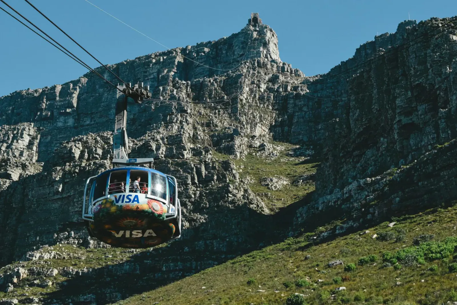 Visa-branded cable car ascending steep cliffs of Table Mountain in Cape Town under blue sky.