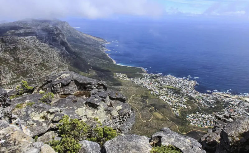 Panoramic view from Table Mountain ridge over Cape Town, ocean, and city below under partly cloudy skies.