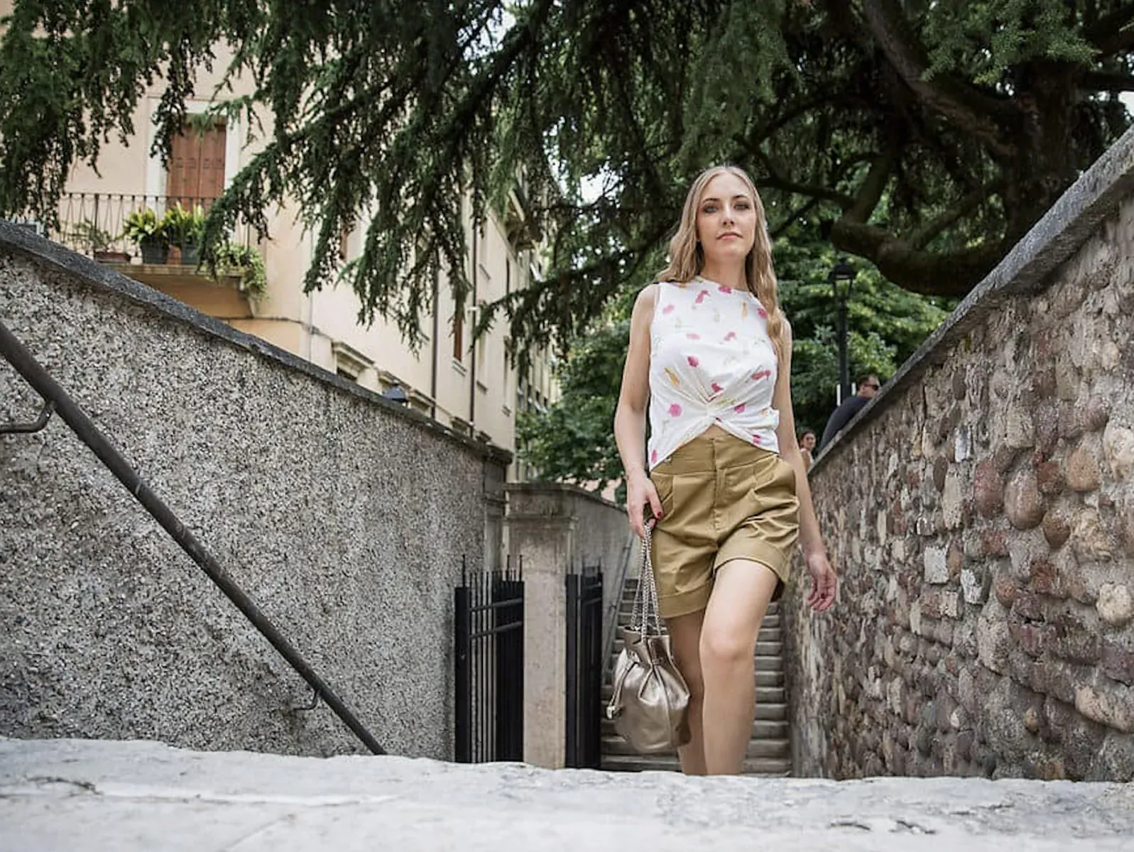 Blonde woman in white floral tank top and khaki shorts with gold bag walks up stone steps in Italian street amid trees