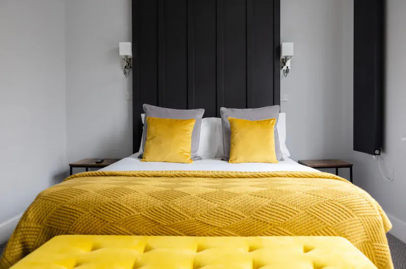 Cozy hotel bed with black headboard, yellow pillows and textured throw, flanked by wall lamps and nightstands.