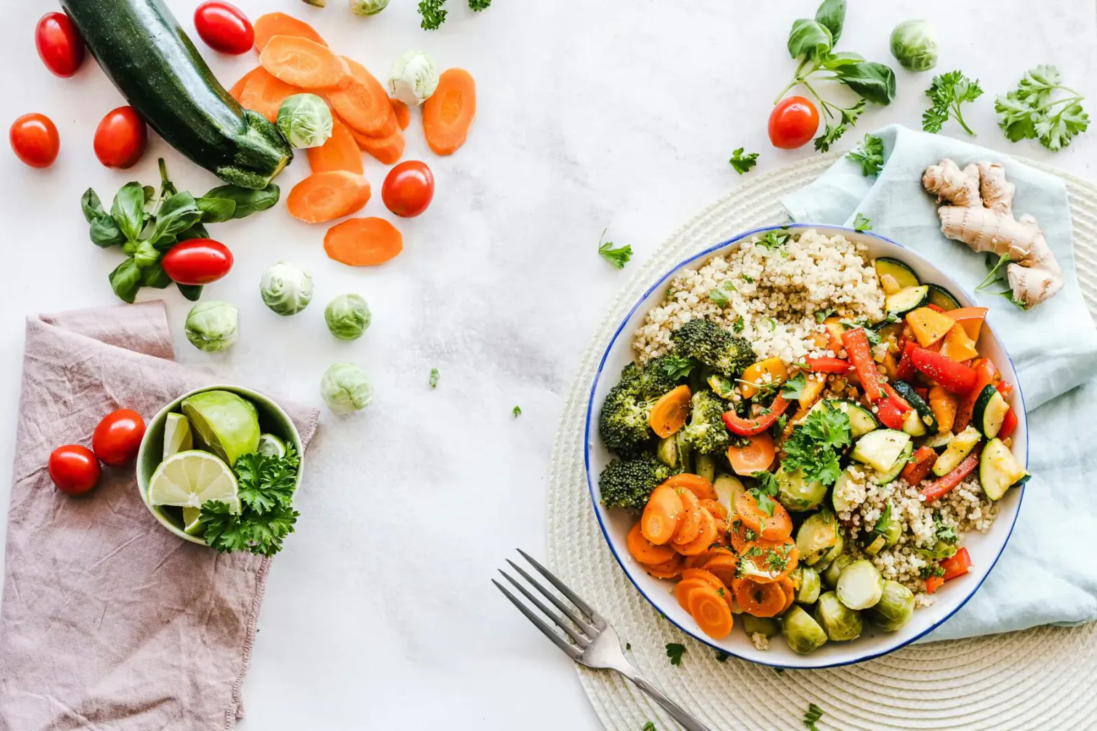 Vibrant vegetable stir-fry with quinoa in blue bowl, surrounded by fresh carrots, zucchini, broccoli, tomatoes, lime, ginger on white table.
