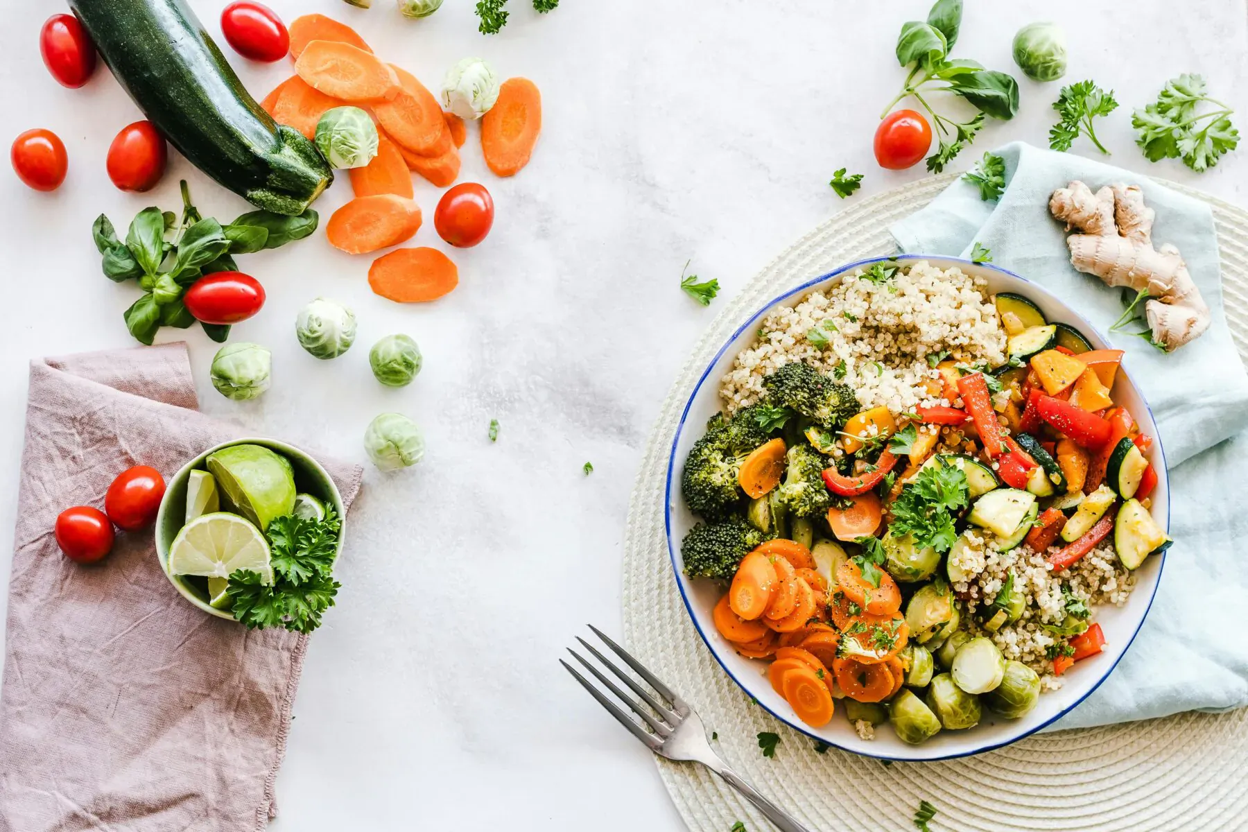 Vibrant vegetable stir-fry with quinoa in blue bowl, surrounded by fresh carrots, zucchini, broccoli, tomatoes, lime, ginger on white table.