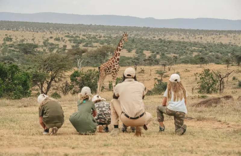 Family in safari gear kneeling excitedly near a tall giraffe in African savanna with acacia trees.