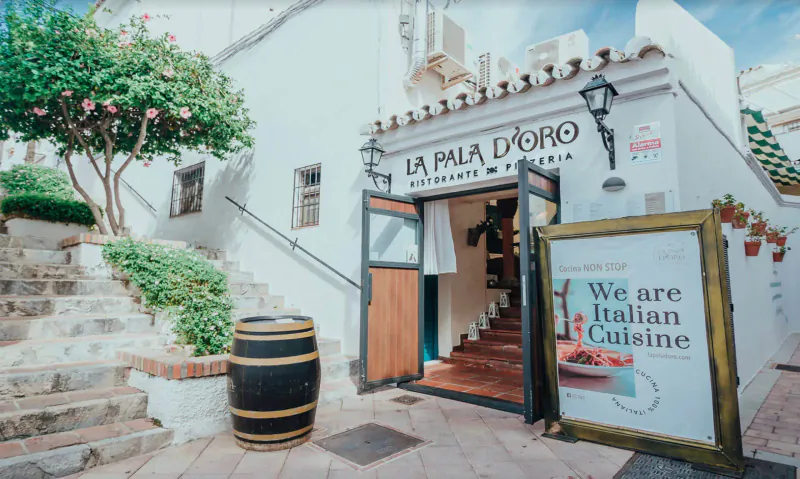 La Pala d'Oro Italian restaurant exterior on Costa del Sol with 'We are Italiani Cuisine' sign, plants, barrel, and steps.