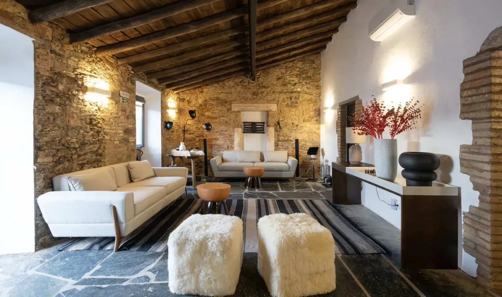 Rustic stone-walled living room with wooden beams, white sofas, ottomans, red flowers, and modern decor.