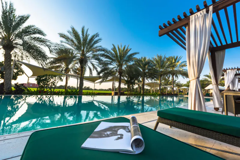 Luxury poolside lounge chair with open magazine at Melia Desert Palm Dubai resort, palm trees and blue sky.