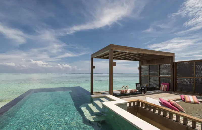 Luxury overwater villa in Maldives with infinity pool overlooking turquoise lagoon, pink cushions on deck chairs under wooden pergola.